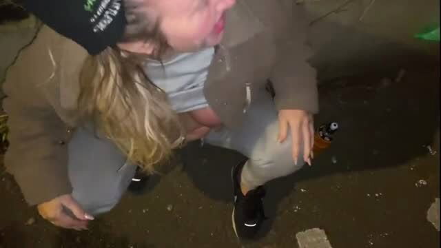 Blonde woman kneeling outdoors, mouth open, alcohol bottle on ground
